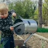 A young boy standing beside a watering can in a garden